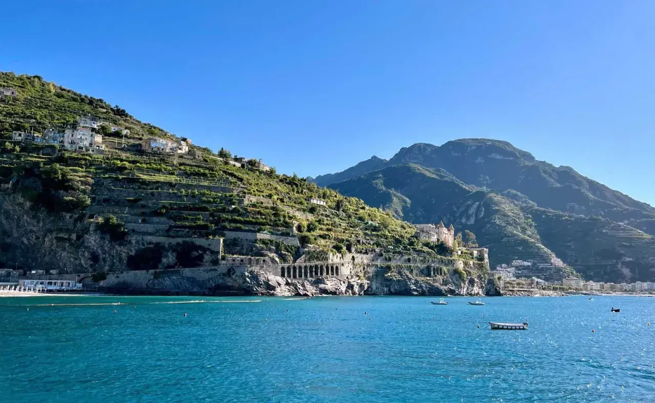 A scenic view from a Naples to Amalfi Coast ferry, showing terraced hillsides dotted with houses, a grand villa, and the sparkling blue sea.