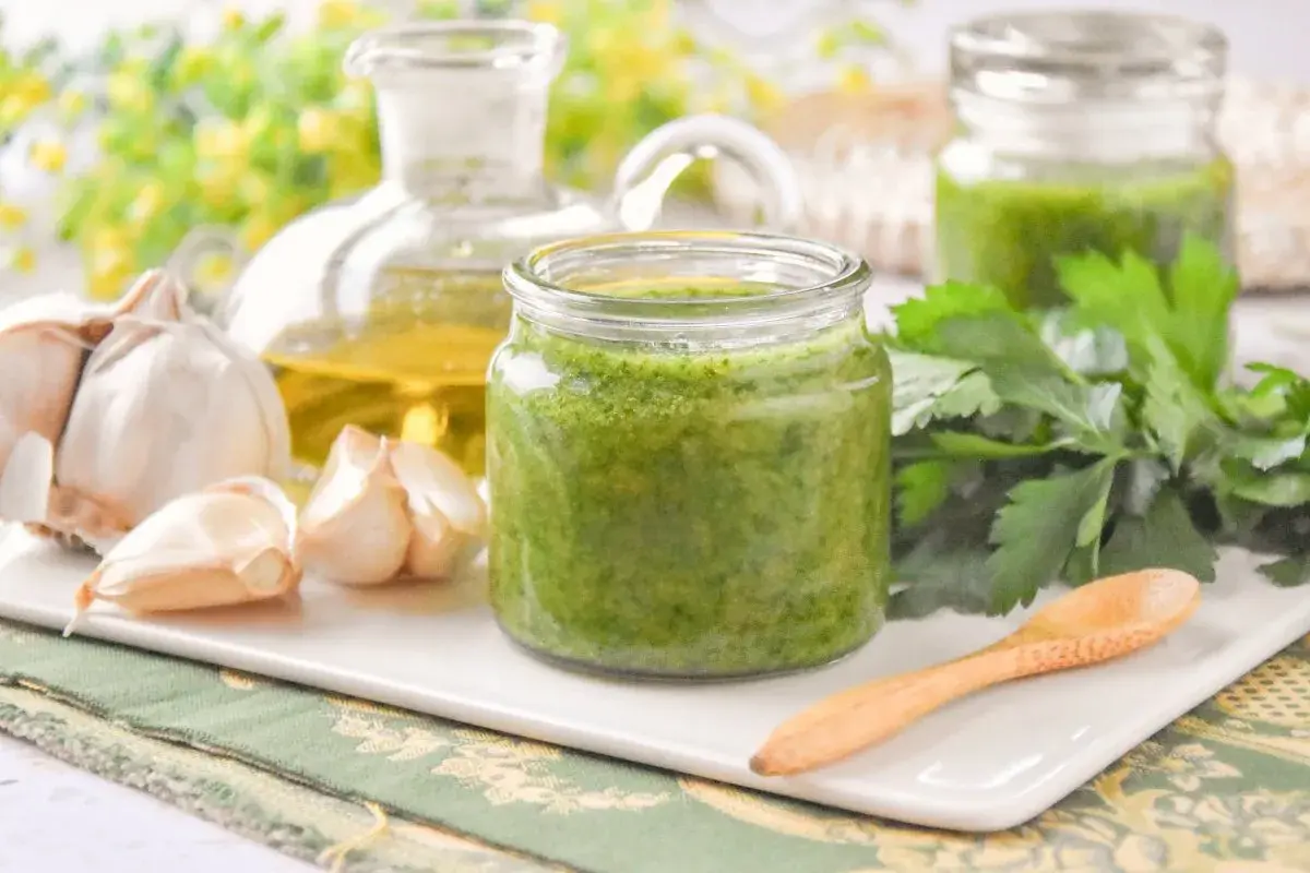 Un frasco de salsa mery verde vibrante, con ajo, aceite de oliva y perejil fresco alrededor.