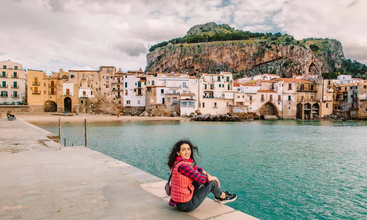 A woman smiles, enjoying her first time visiting Sicily. She sits on a stone pier overlooking the turquoise sea and a picturesque town with a dramatic cliff behind it.