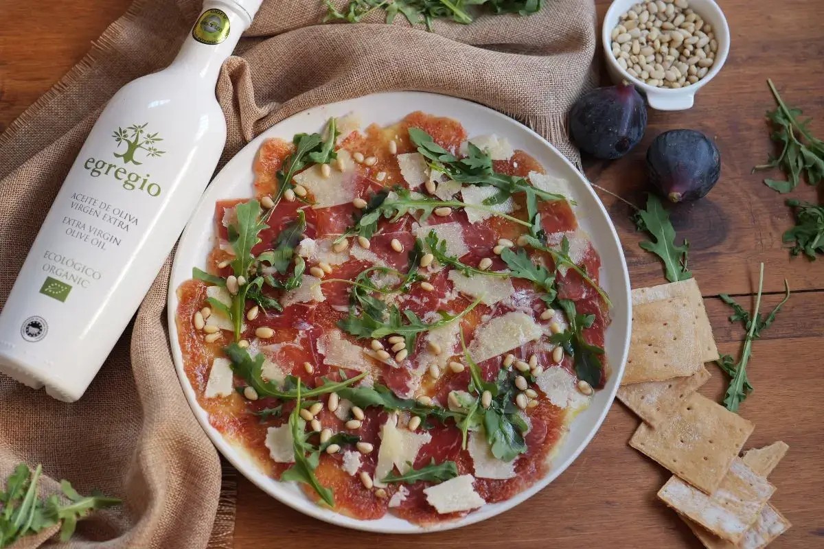 Delicioso carpaccio de higos con jamón serrano, piñones, rúcula y lascas de queso. Un plato ligero y lleno de sabor.