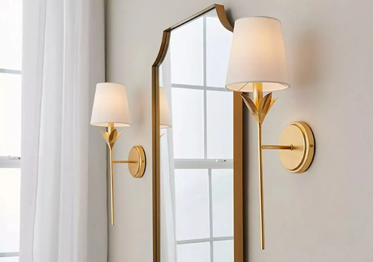 Apliques de pared dorados con pantalla blanca, junto a un espejo con marco dorado.