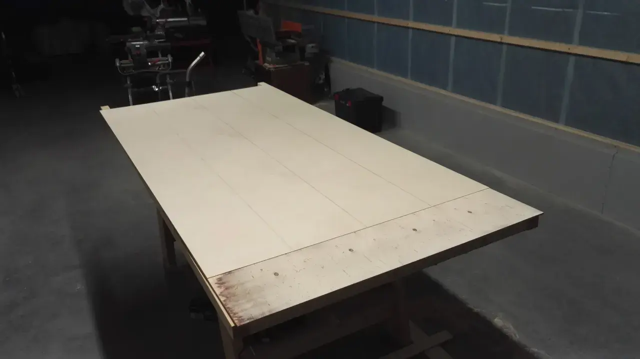 Stor skiva av plywood på ett arbetsbord, redo att bygga slagport. Verktyg syns i bakgrunden.