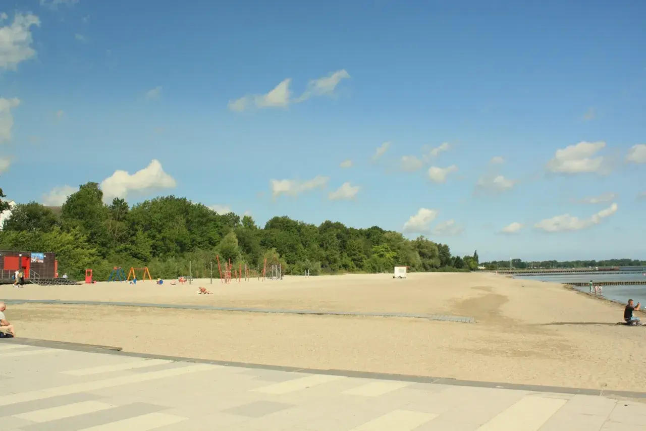 Sonniger **Stralsund Strand** mit Spielplatz, Liegewiese und Blick aufs Wasser. Ein Mann macht ein Foto.