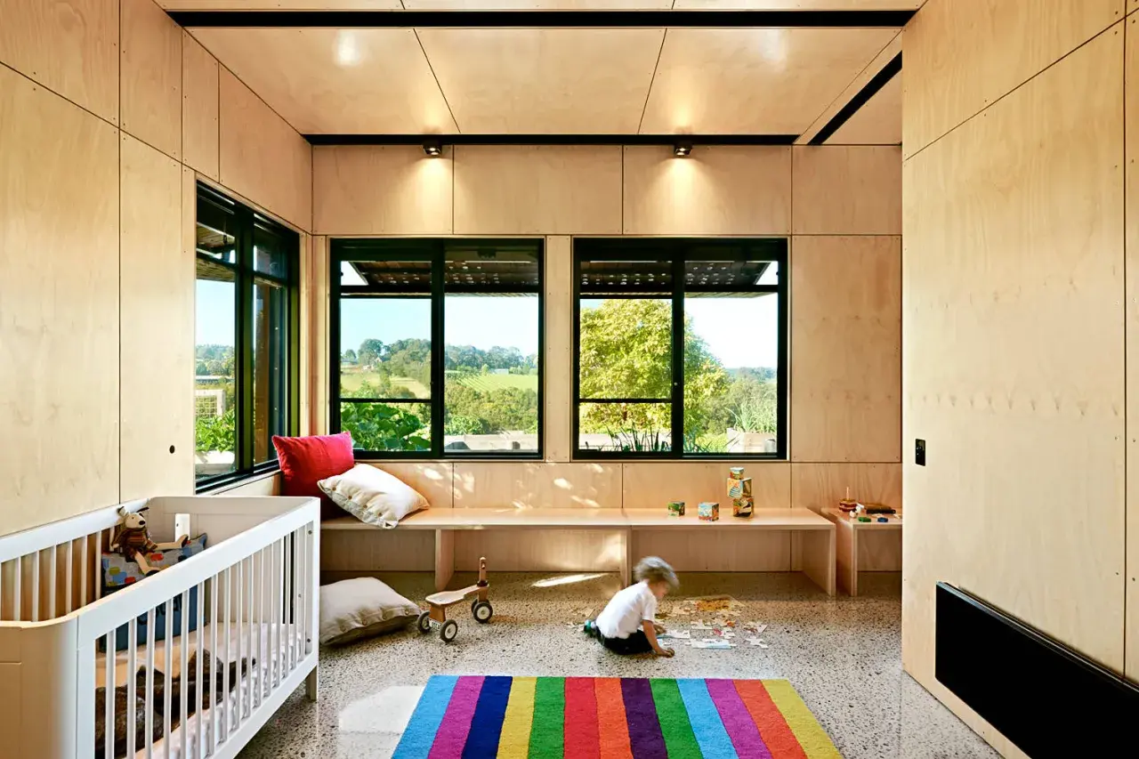 Barnrum med väggar och tak av plywood som ytskikt. Ett barn leker med pussel på golvet framför en bänk vid fönstret.