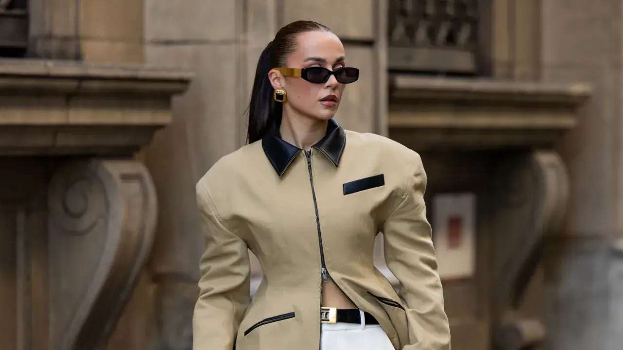 Mujer con gafas de sol oscuras y pendientes dorados, vistiendo una chaqueta beige con cuello negro y cinturón. Un look muy chic.