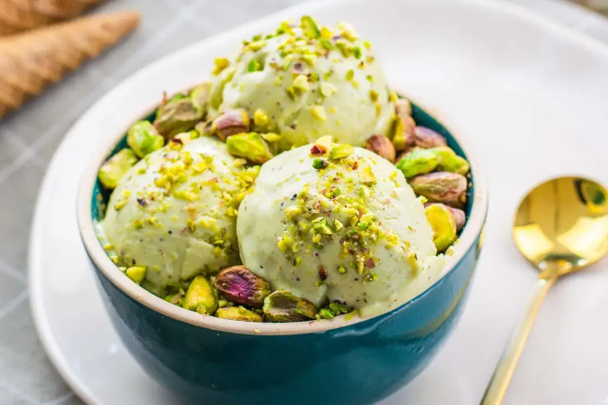 Tre palline di cremoso gelato al pistacchio, guarnite con granella e pistacchi interi, in una ciotola blu.