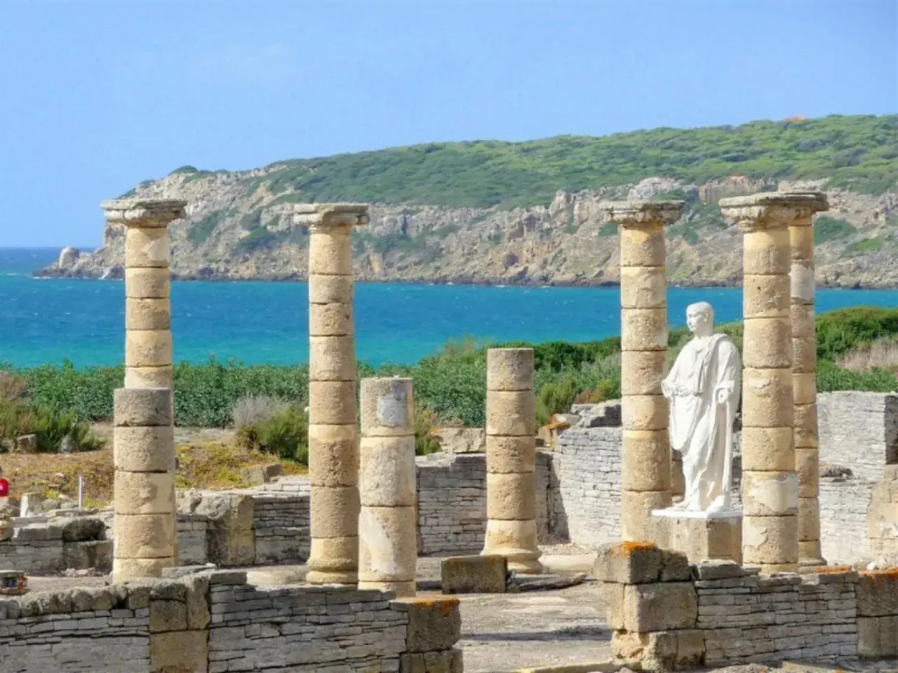 Ruinas romanas en el área de Bolonia, con columnas de piedra y una estatua blanca frente al mar azul y una colina verde.