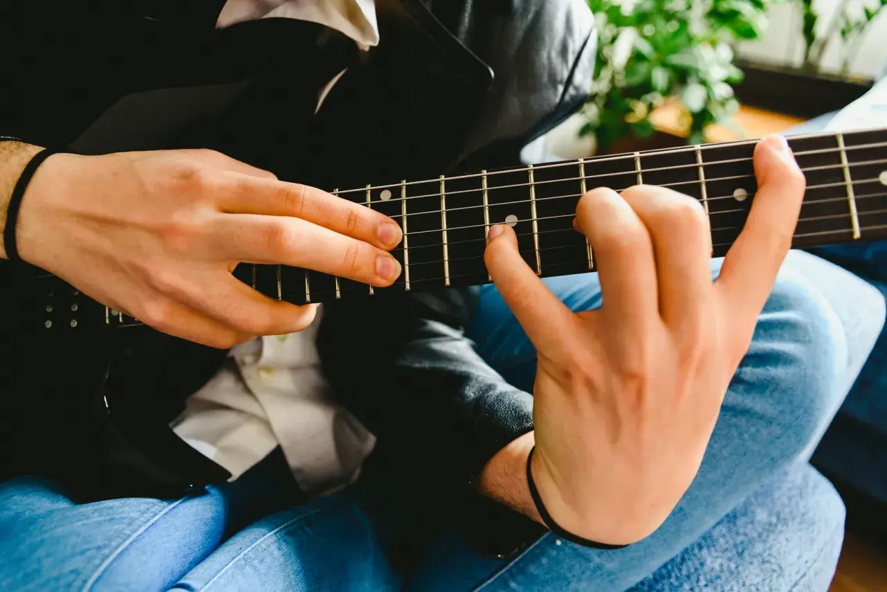 Dłonie grające na czarnej gitarze elektrycznej, z widocznymi progami i strunami.