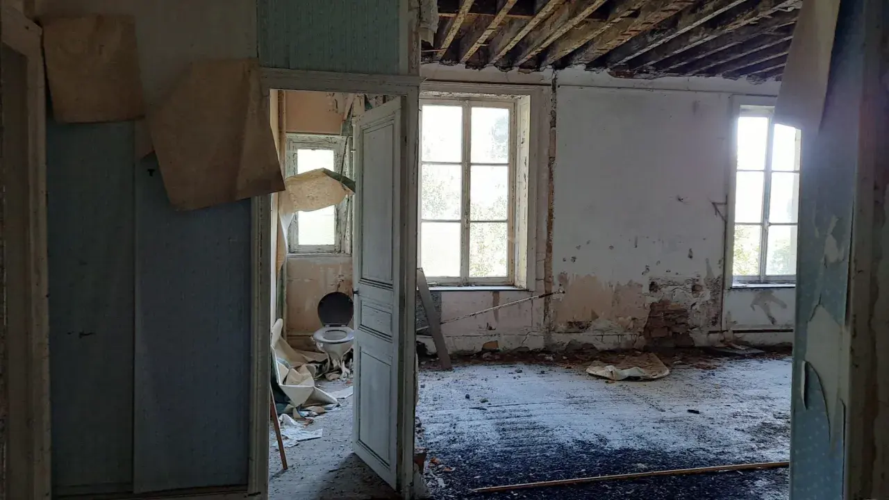 Intérieur délabré d'une maison abandonnée dans les Ardennes, avec un papier peint déchiré, un plafond effondré et une vieille cuvette de toilette. Urbex Ardennes.