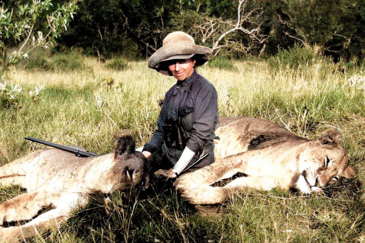 En kvinna med hatt och kikare sitter bland två lejon. Hon påminner om Karen Blixens alter ego, redo för äventyr i Afrika.