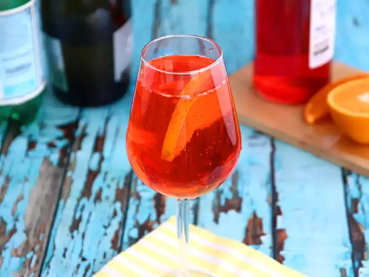 En Aperol Spritz, Italiens nationaldryck, med apelsinskiva i glaset. Bakom syns flaskor och en apelsinhalva.