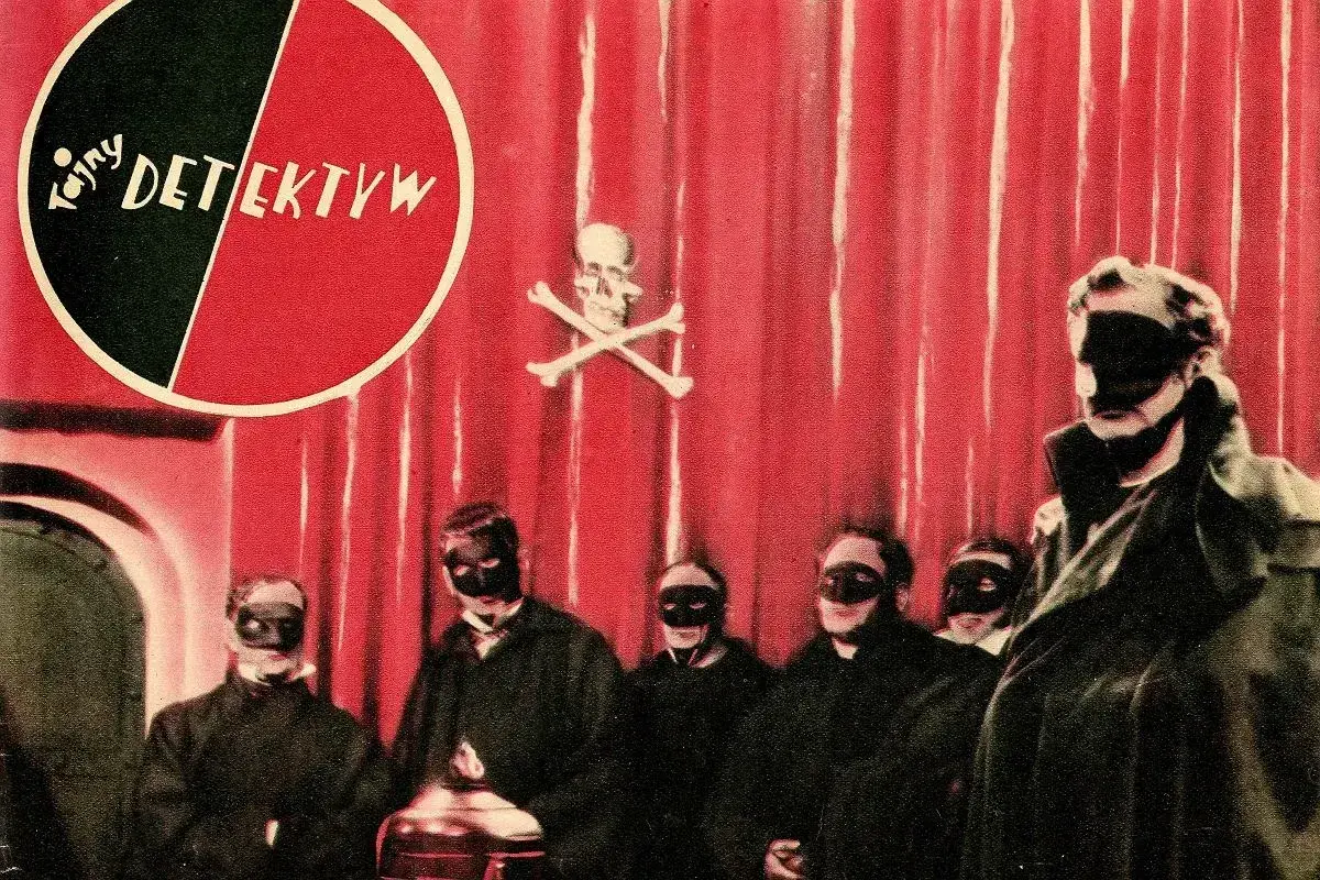 Grupa zamaskowanych postaci w czarnych strojach, na czerwonym tle z symbolem czaszki i piszczeli. Może to być scena z tajemniczej czarnej mszy.