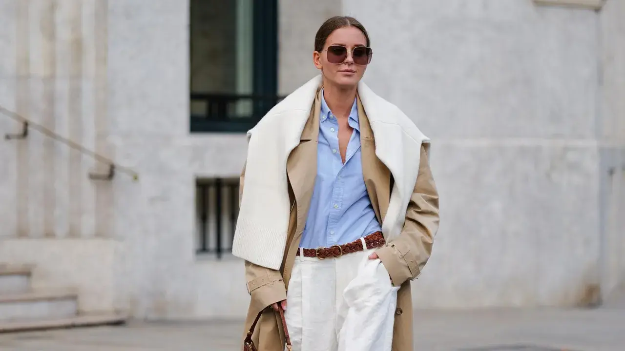 Eine Frau trägt einen beigefarbenen Trenchcoat, ein hellblaues Hemd und weiße Hose. Das Outfit zeigt, wie man ein beiges Oberteil kombinieren kann, auch für Herren.