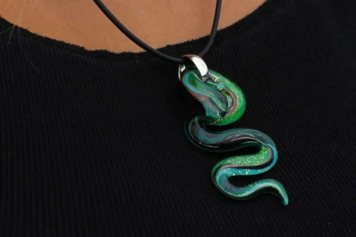 Un serpente sinuoso de **colgante de cristal de Murano** en tonos verdes y azules, con destellos dorados, descansa sobre una tela negra.