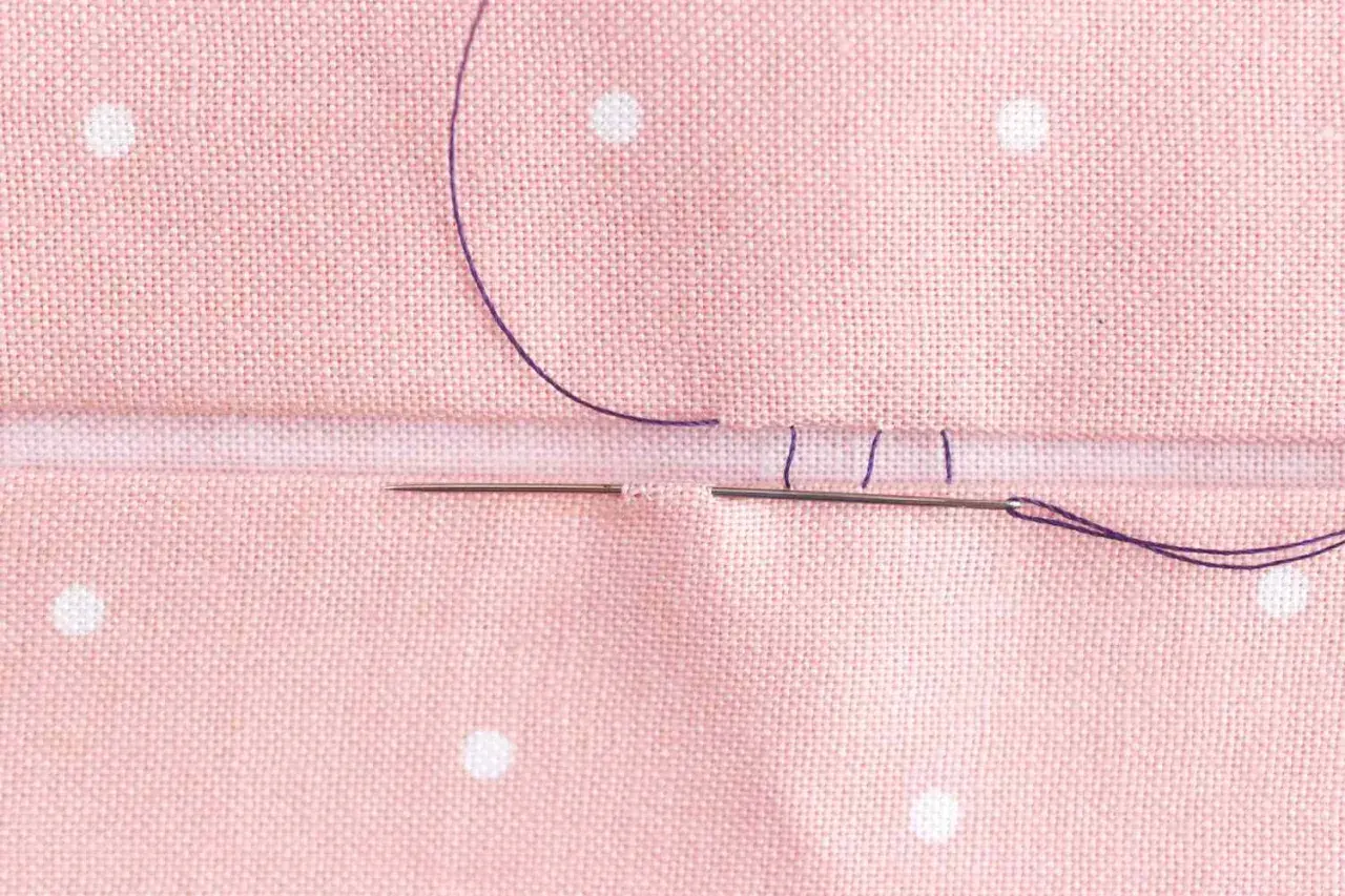Close-up of a needle and purple thread creating an invisible hem stitch on pink polka-dot fabric.