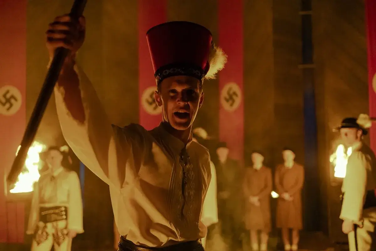 Młody mężczyzna w czerwonym nakryciu głowy z piórem wznosi płonący kij. W tle widać flagi ze swastykami, sugerujące scenę z filmu "Biała odwaga".