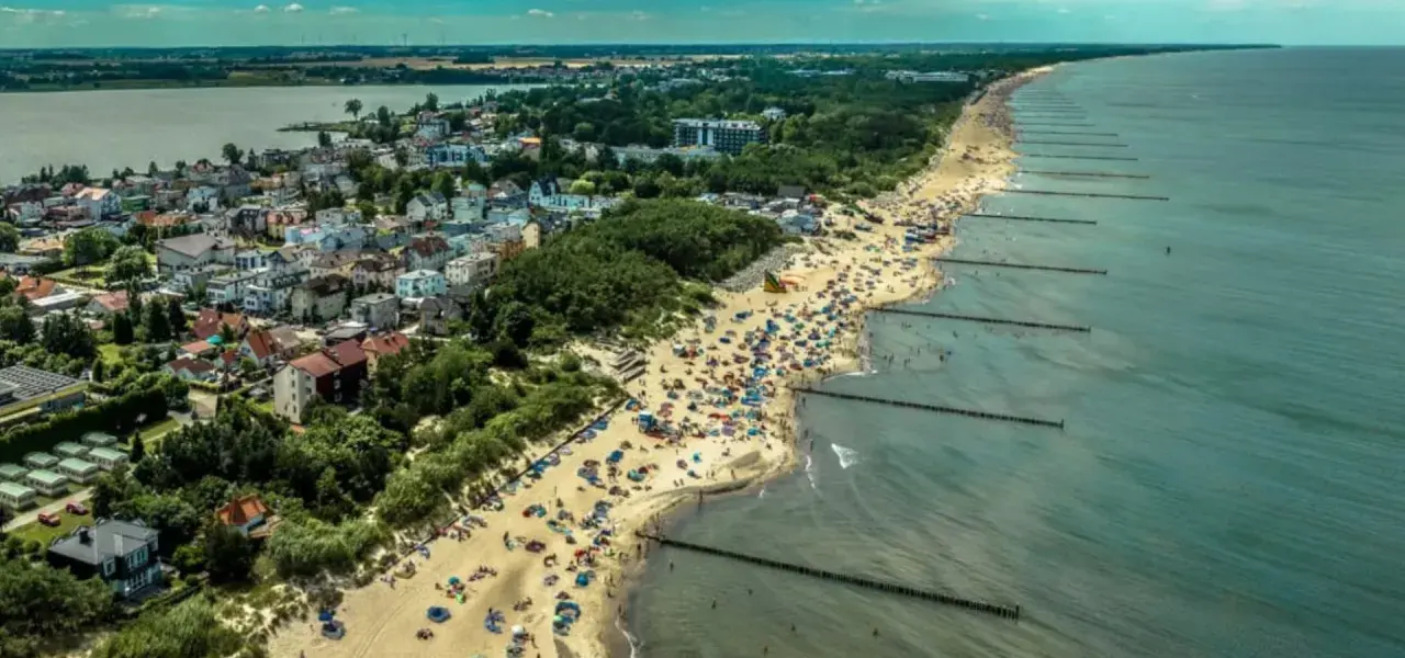 Tłum ludzi na plaży w Mielnie, cieszących się słońcem i morzem. W tle widać zabudowania i jezioro. To idealne miejsce na wakacje, pełne atrakcji.