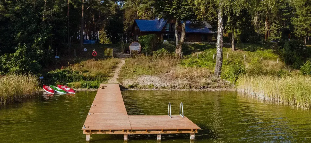 Drewniany pomost nad jeziorem w Chojnicach, obok trzy kajaki. W tle dom wśród drzew.