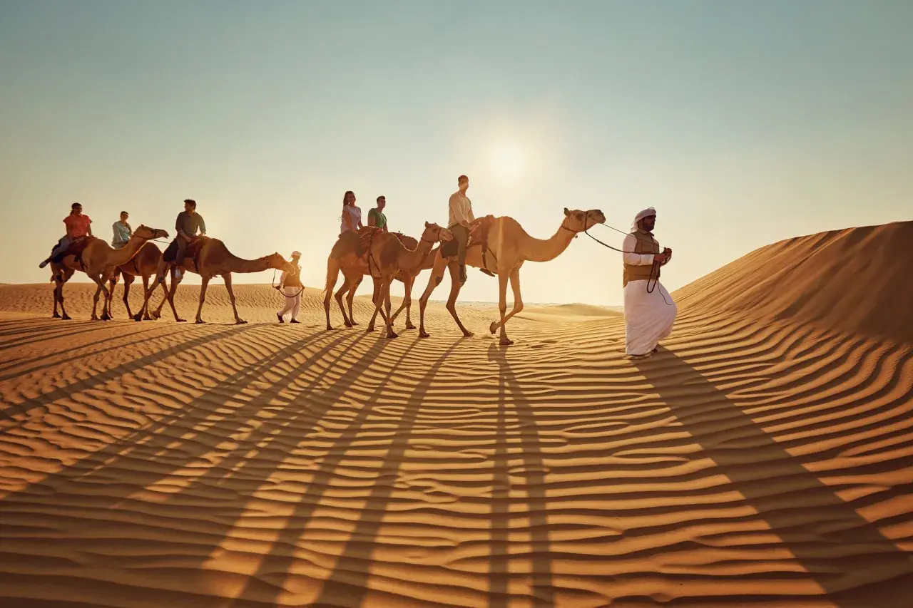 Kamelkarawane in der Wüste bei Sonnenuntergang. Ein Führer in traditioneller Kleidung begleitet Touristen. Tolle Abu Dhabi Tipps für ein unvergessliches Erlebnis.