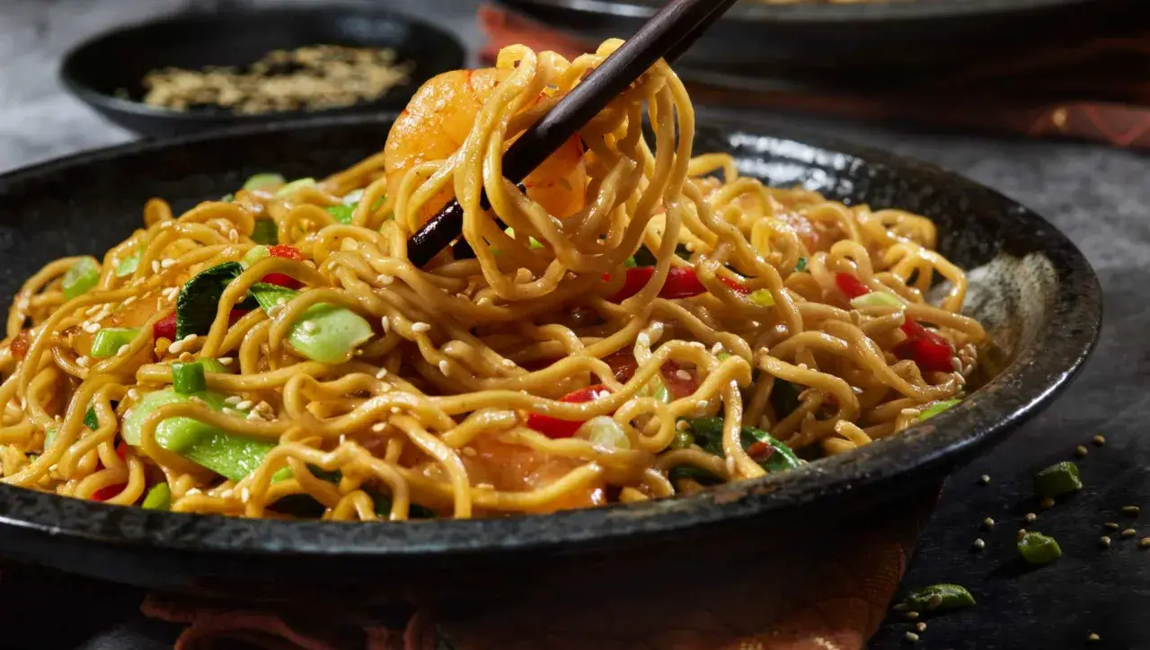 Un plato humeante de fideos con pollo, salteado con verduras y semillas de sésamo, listo para ser disfrutado.