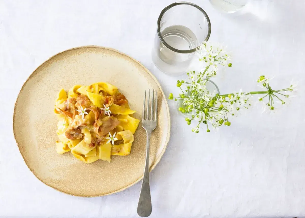 Ein Teller mit cremiger Pasta, die wie Fettuccine aussieht, garniert mit zarten Blüten und Speck. Daneben ein Glas Wasser und ein Strauß weißer Blumen.