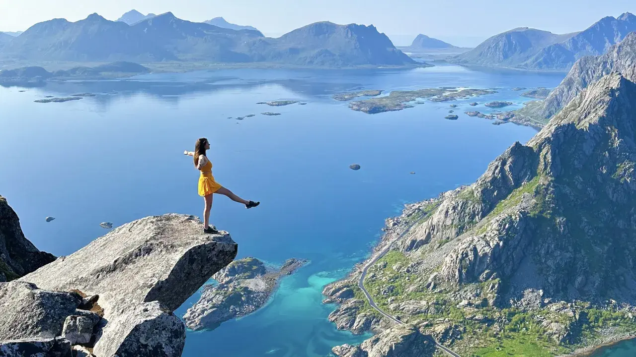 Une femme en robe jaune sur un promontoire rocheux, admirant les plus beaux endroits des îles Lofoten, avec des montagnes bleues et des eaux turquoise.