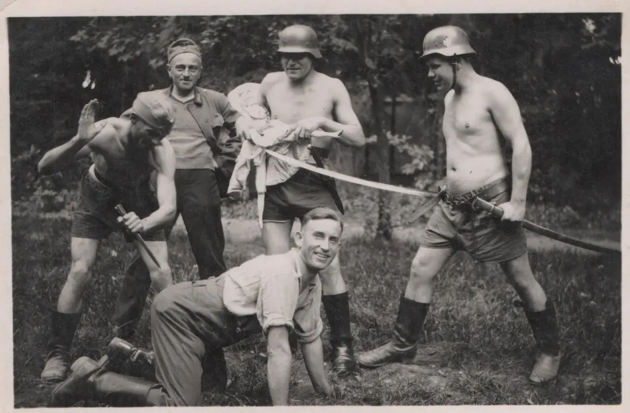 Fünf Männer, einige mit Helmen, posieren mit Waffen. Ein Mann hält eine Puppe. Die Szene erinnert an die Verbrechen der Wehrmacht.
