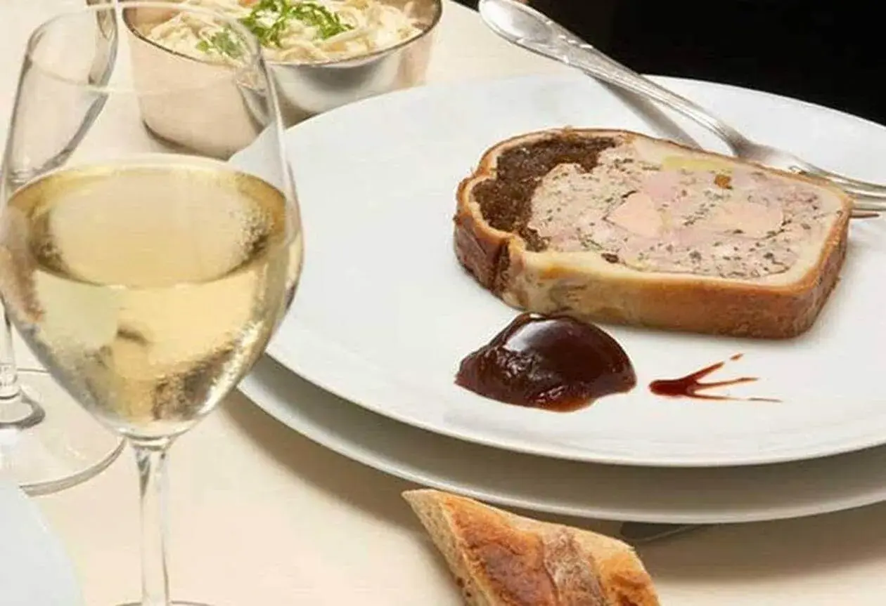 Une tranche de pâté en croûte, garnie de confiture de figues, accompagnée d'un verre de vin blanc et d'une coupelle de crudités.