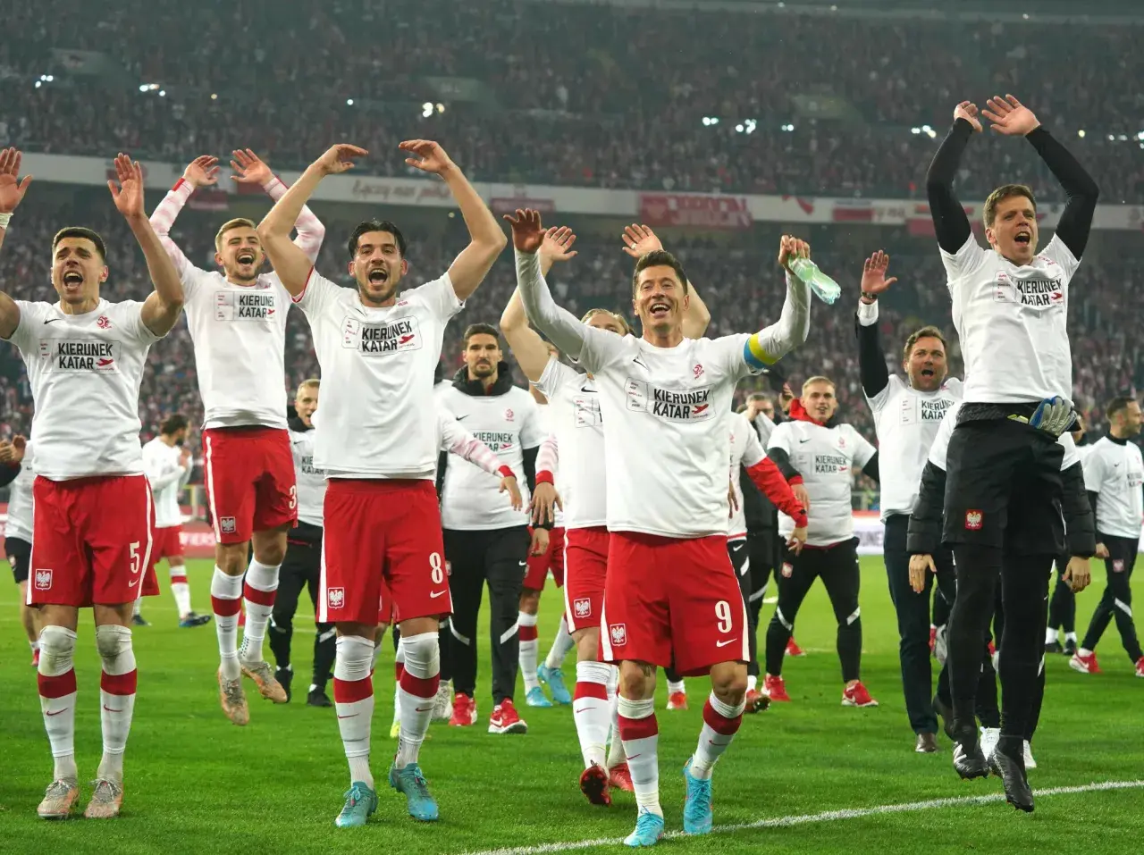 Piłkarze reprezentacji Polski w białych koszulkach i czerwonych spodenkach świętują na boisku.
