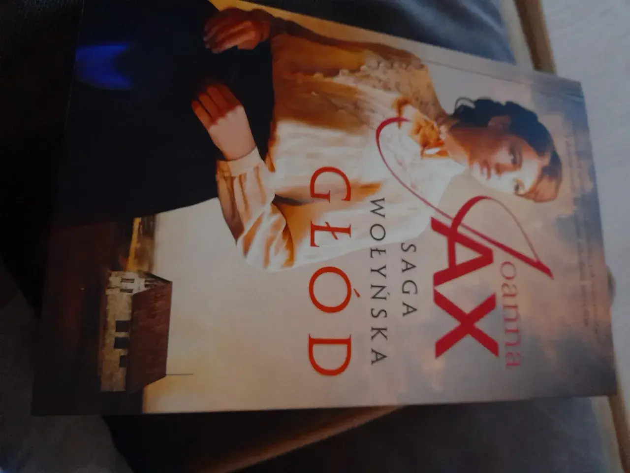 Okładka książki "Głód. Saga Wołyńska" Joanny Jax z portretem kobiety i budynkiem.