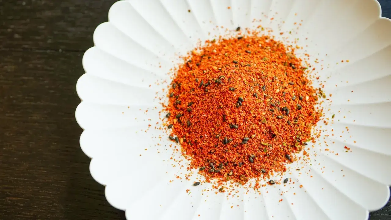 A pile of vibrant Japanese 7 spice blend, featuring chili flakes and black sesame seeds, sits on a white, fluted dish.