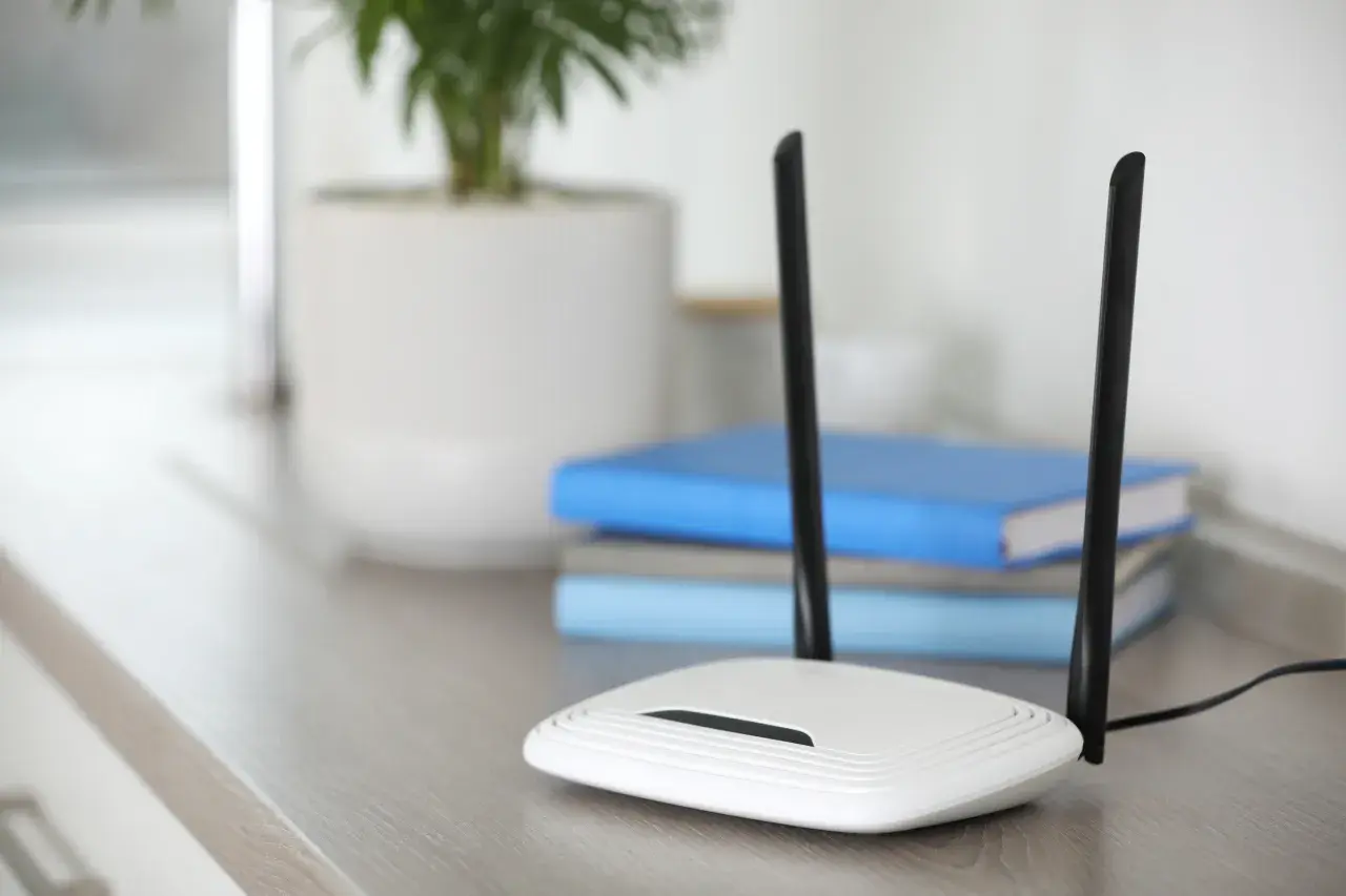 Biały router z dwiema antenami, zapewniający szybkie **standardy wifi**. W tle książki i roślina w doniczce.