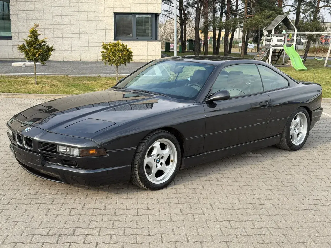 Czarne BMW V12, klasyk z lat 90., zaparkowane na kostce brukowej przed budynkiem z placem zabaw w tle.