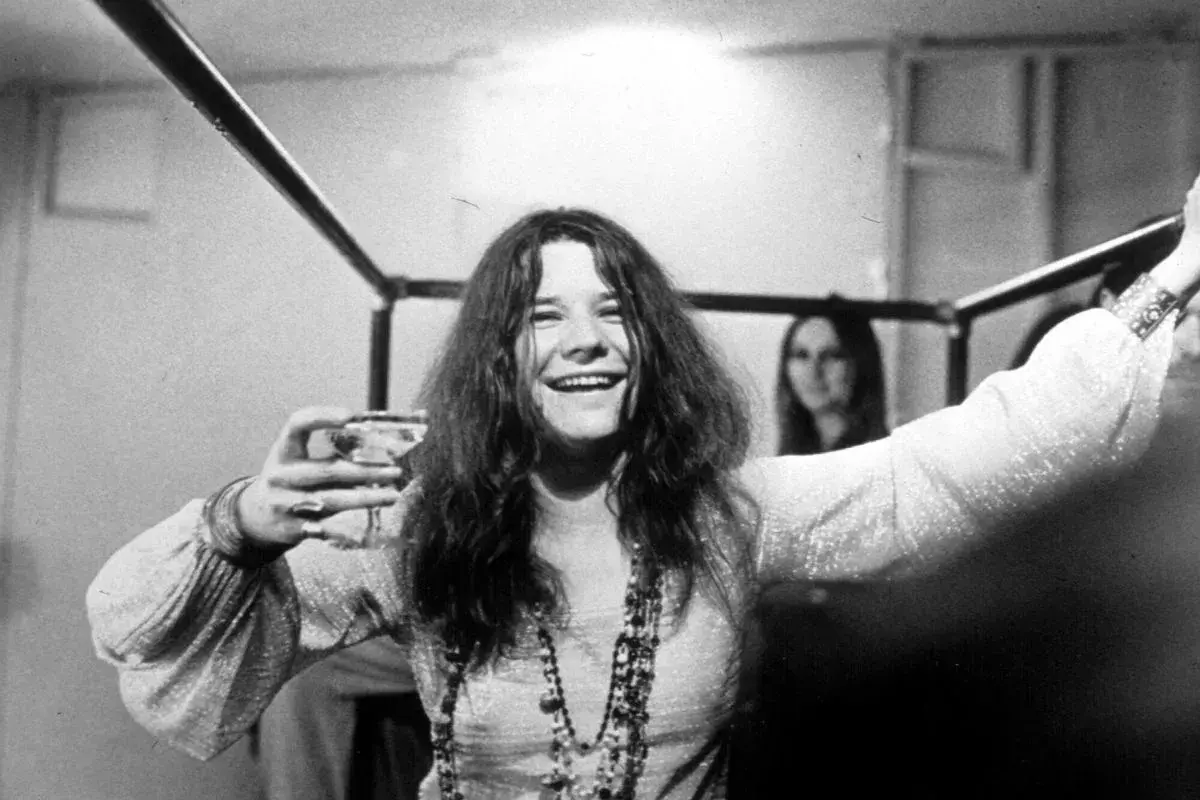 Janis Joplin, z kieliszkiem w dłoni, uśmiecha się szeroko. Jej charakterystyczne włosy opadają na ramiona, a na szyi wisi naszyjnik. W tle widać inną kobietę. To zdjęcie przypomina o jej niezapomnianych **janis joplin utworach**.