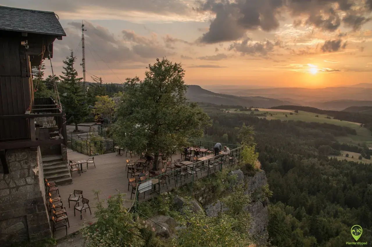 Widok z tarasu restauracji na Górach Stołowych o zachodzie słońca. Drewniany taras z meblami, otoczony skalistym zboczem i lasem.