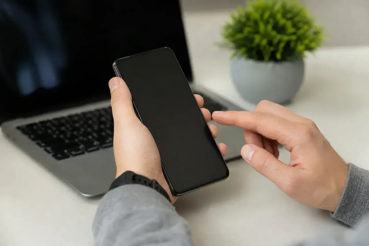 Dłoń trzyma smartfon, palec wskazujący dotyka ekranu, przygotowując się do całkowitego czyszczenia telefonu. Obok laptop i roślina.