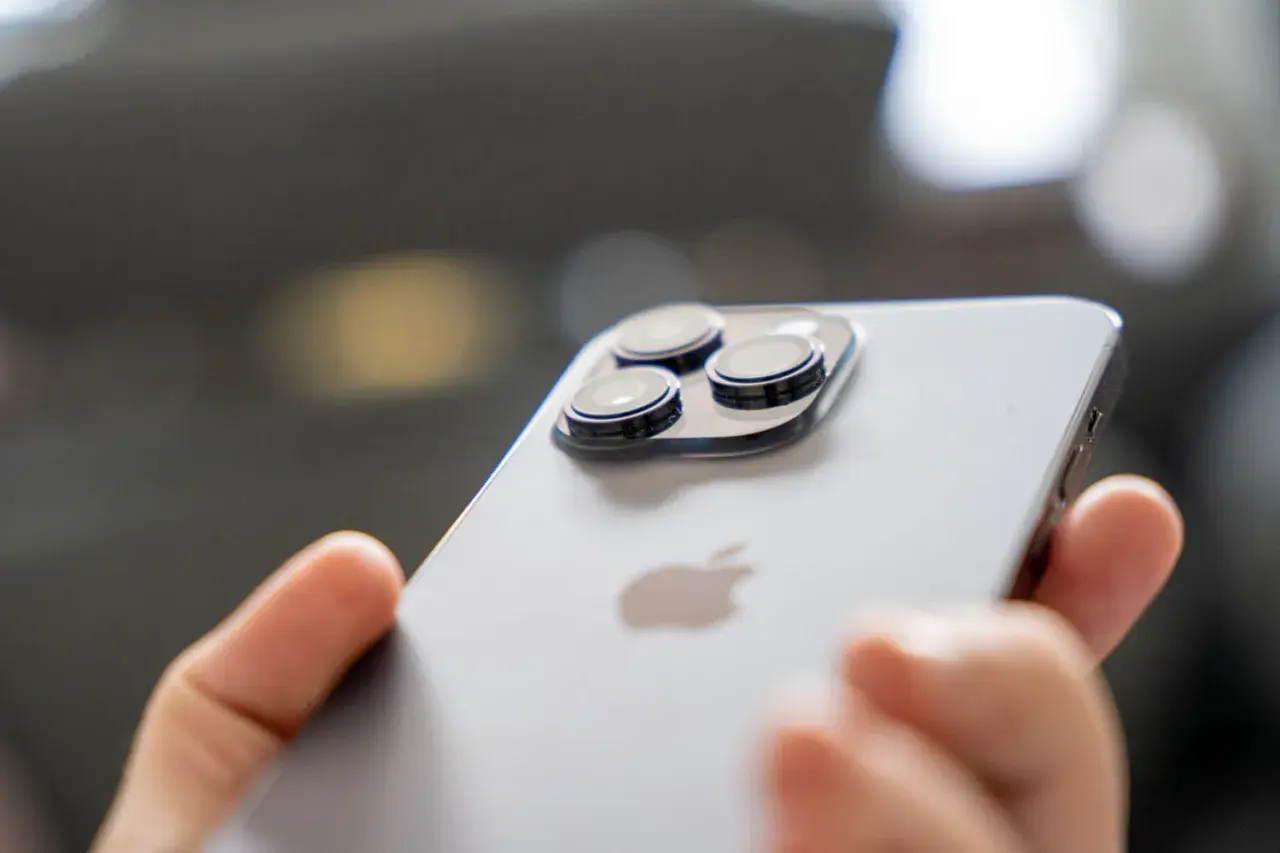 Dłoń trzyma iPhone 14 Pro, pokazując jego potrójny aparat. Długi czas pracy na baterii pozwala na swobodne korzystanie.