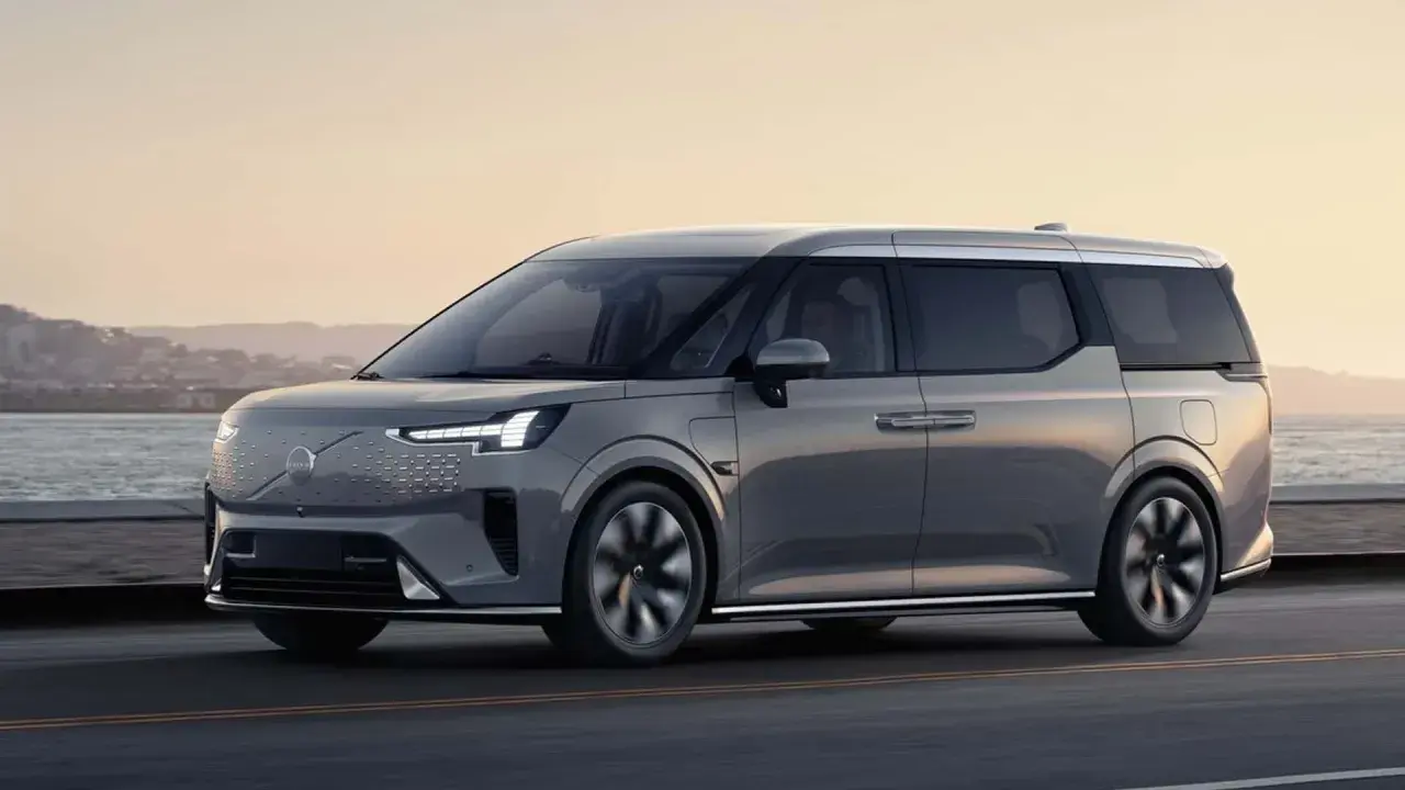 Elegancki, szary minivan Volvo EM90 jedzie wzdłuż wybrzeża o zachodzie słońca.