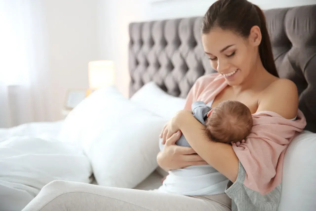 Uśmiechnięta mama karmi piersią noworodka. Czas karmienia jest elastyczny, ważne by dziecko było najedzone.