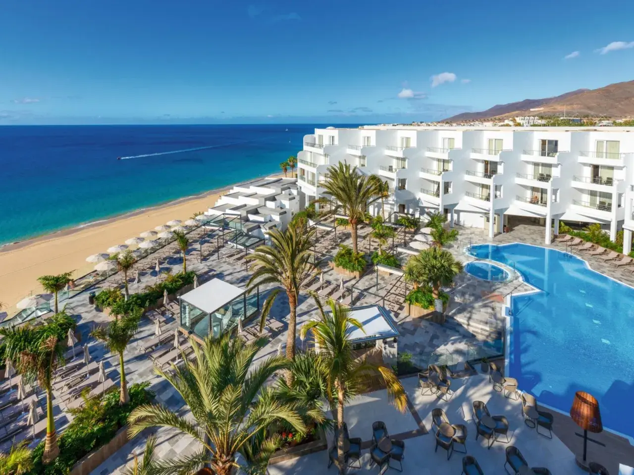 Traumhafter Blick auf das Riu Palace Jandia: Weißes Hotel, Pool, Palmen und der goldene Strand am türkisfarbenen Meer.