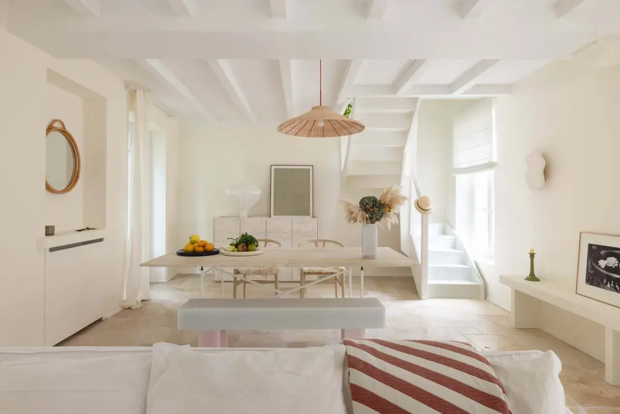 Un soggiorno luminoso con soffitto a travi e **bianco caldo pareti**. Tavolo da pranzo in legno, sedie, panca e una scala bianca conducono a un piano superiore.