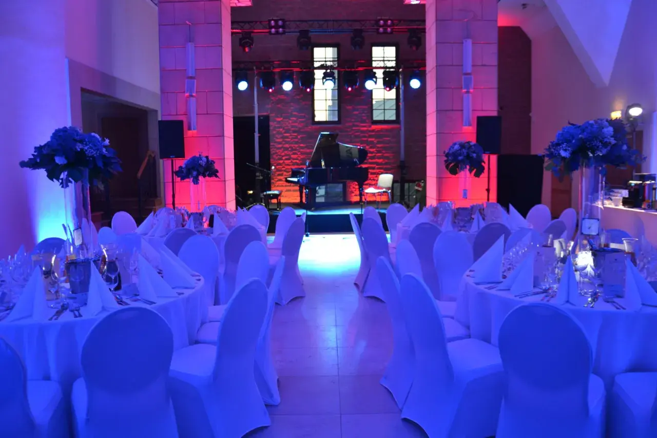 Elegancka filharmonia restauracja z fortepianem na scenie, stoły nakryte białymi obrusami i ozdobione kwiatami, oświetlone niebieskim i różowym światłem.