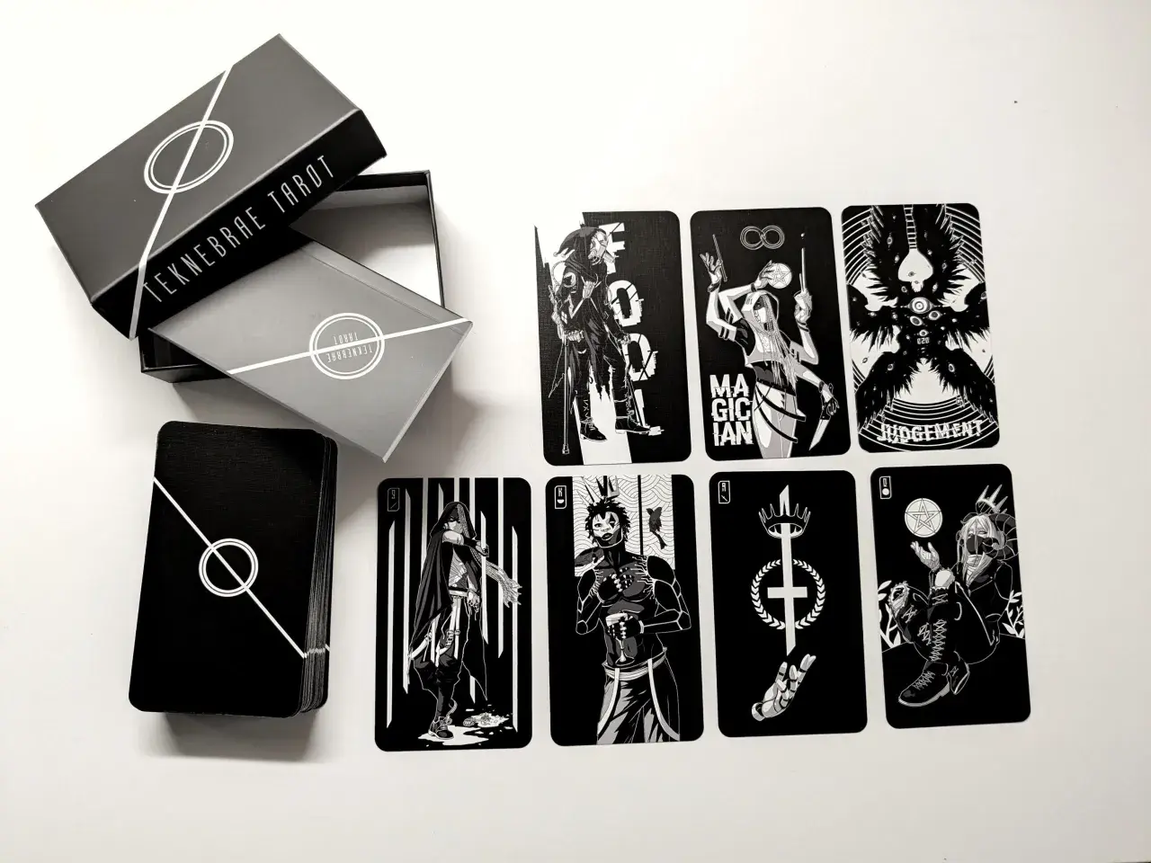 Czarno-białe karty z talii tarot z 5 kartami, w tym Mag, Sąd i inne postacie. Pudełko z napisem "TEKNEBRAE TAROT".