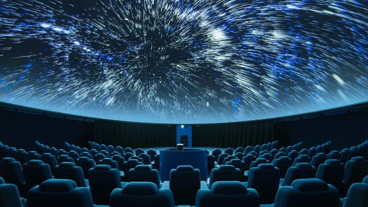 Wnętrze planetarium, gdzie na kopule wyświetlane są rozgwieżdżone niebo i galaktyki. Widownia z niebieskimi fotelami czeka na seans.
