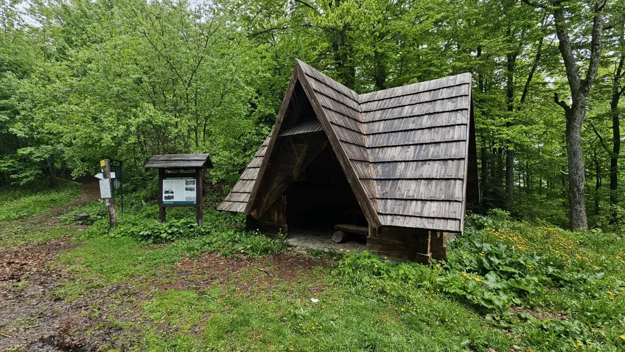 Drewniana wiata na przełęczy żebrak, schronienie pośród zieleni. Obok tablica informacyjna i drogowskaz.