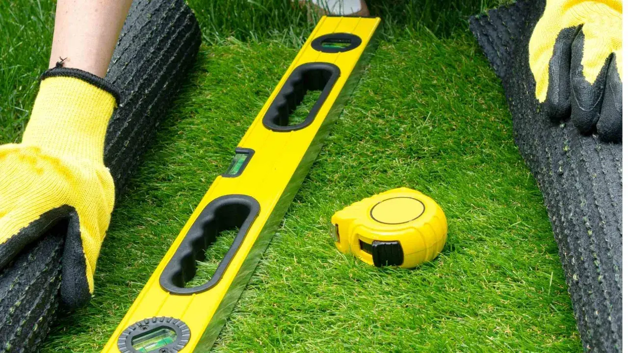 Manos enguantadas preparan terreno para césped artificial. Nivel y cinta métrica listos para el proyecto.