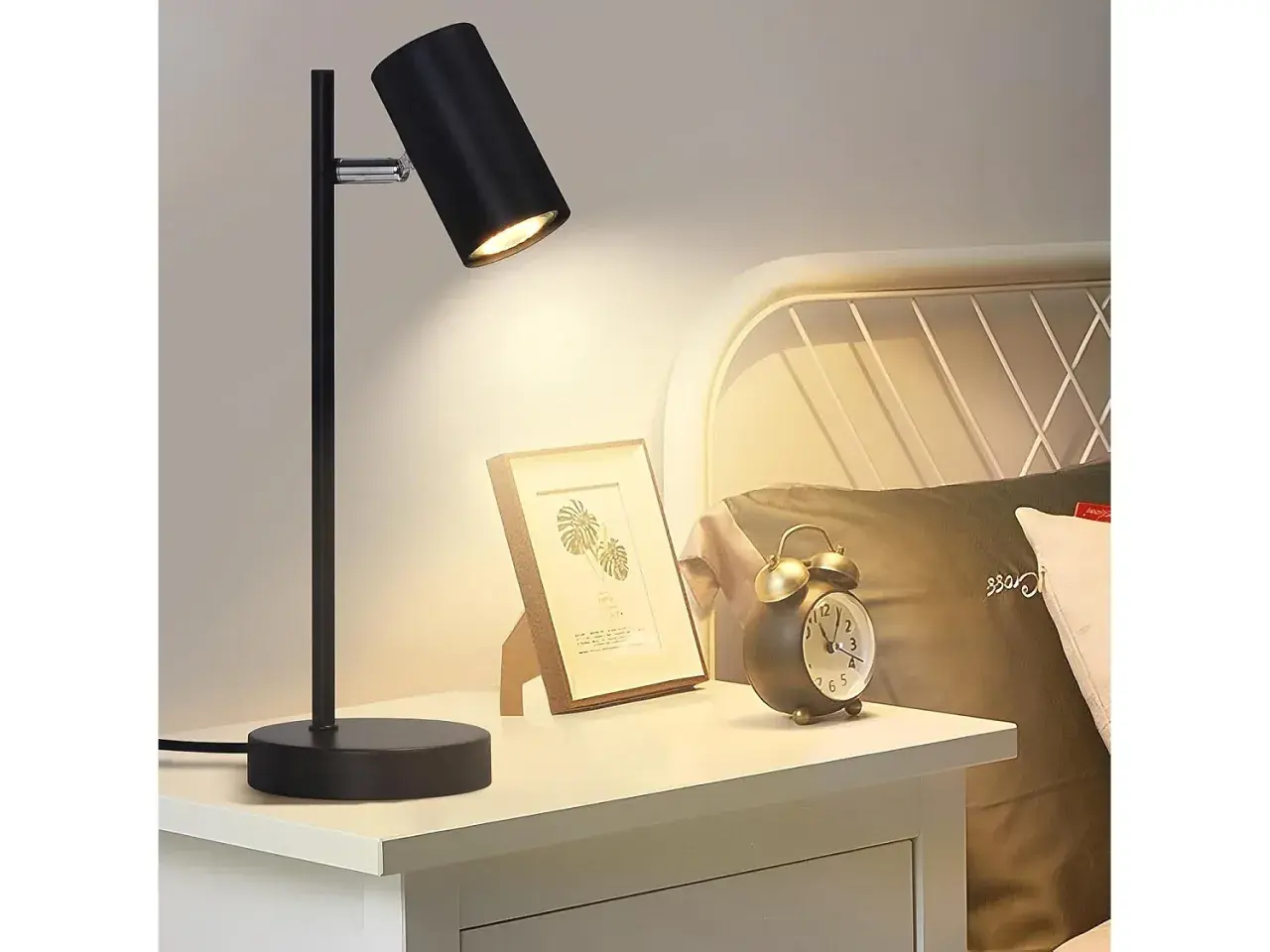 Lampara mesilla negra ilumina un cuadro y un despertador vintage en una mesita de noche junto a una cama.