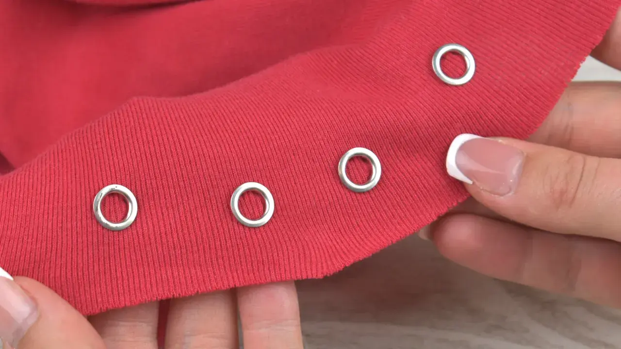 Demonstration of how to put metal rings in fabric. A hand holds red fabric with several silver grommets already attached, and another grommet is about to be placed.