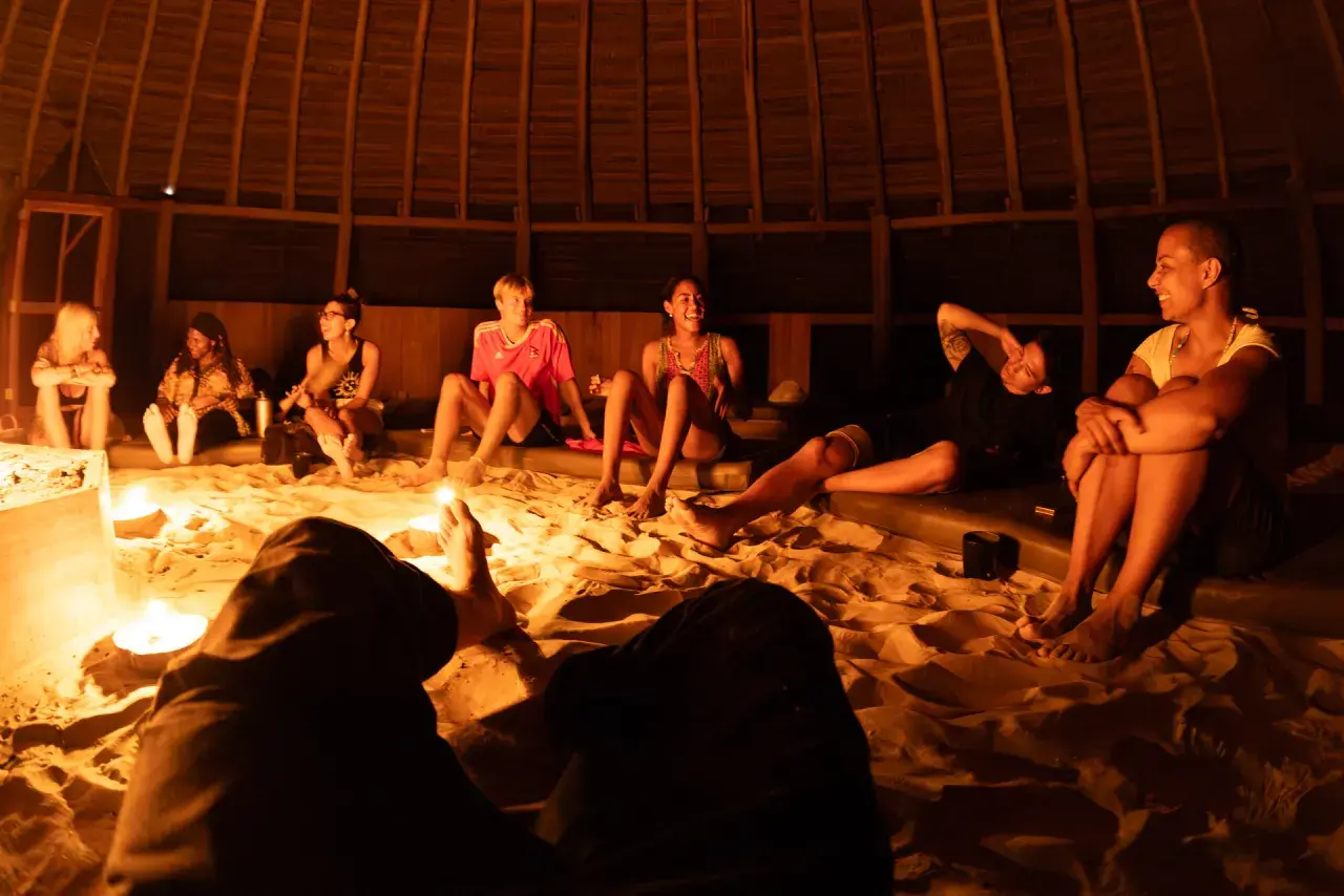 Grupa ludzi siedzi na piasku w okrągłej, drewnianej chacie, otoczona świecami. Rozmawiają i śmieją się, przygotowując się do ceremonii. Ayahuasca cena może być różna, ale to doświadczenie jest bezcenne.