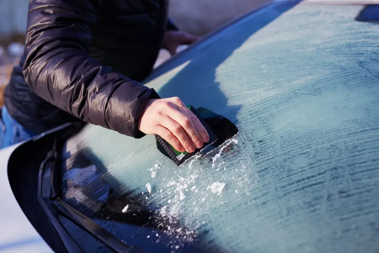 En hand skrapar is från insidan av bilrutan. Snöflingor syns på den frostiga ytan.